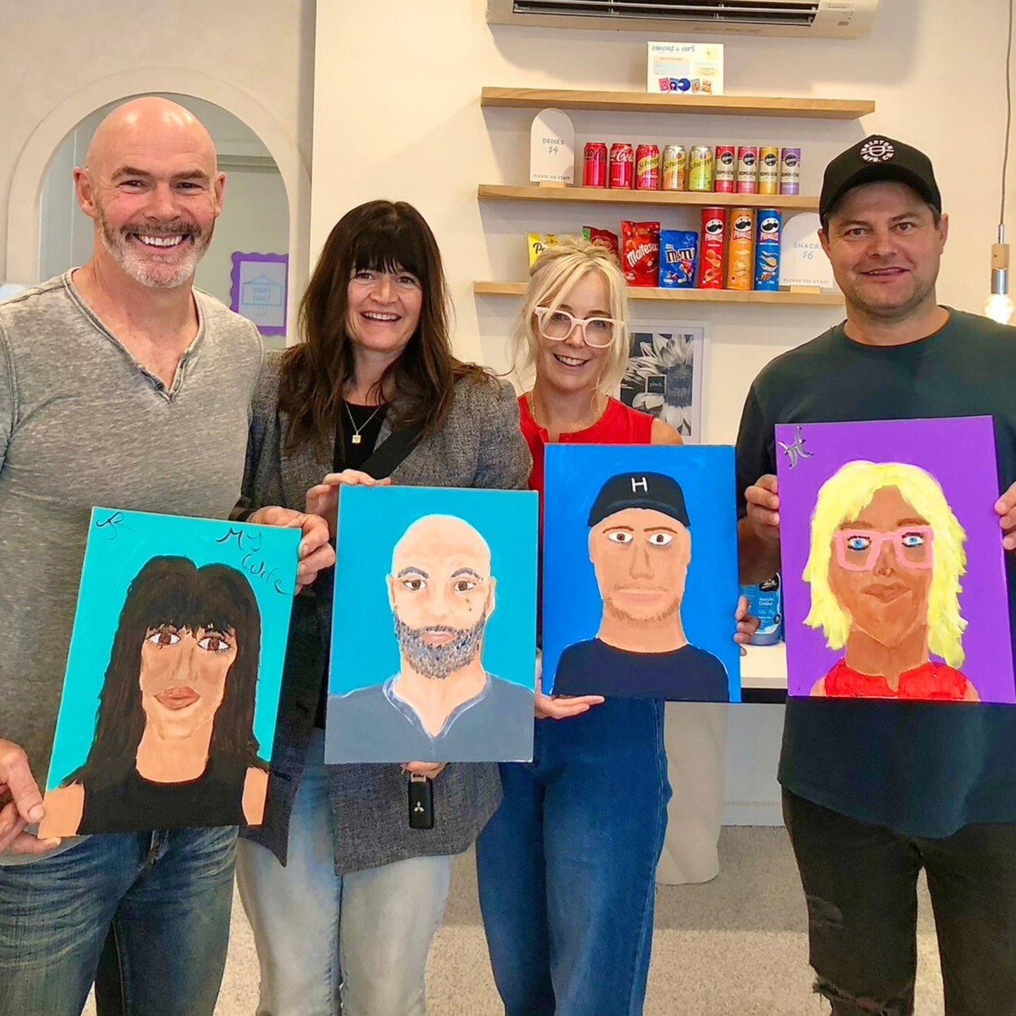 Four people holding up their painted portraits in a room with shelves in the background.